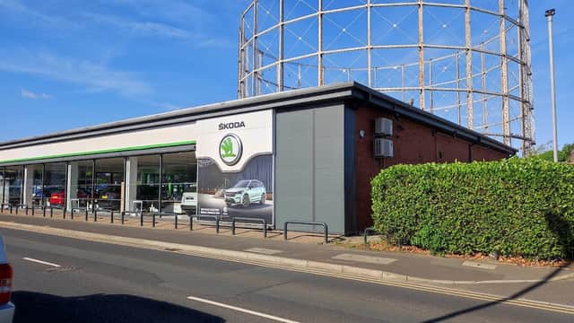 Skoda dealership with a green hedge and a gas holder in the background under a clear blue sky.