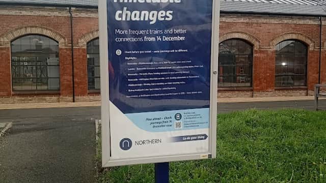 Sign announcing timetable changes for trains, with grassy area and brick building in the background.