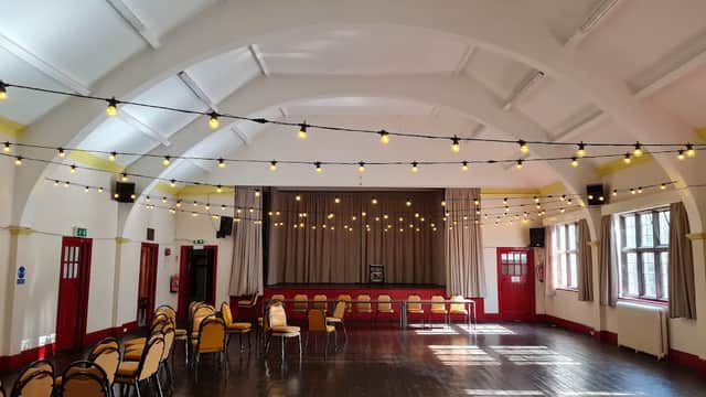 Brightly lit hall with string lights, rows of chairs, and a stage area, featuring large windows and wooden flooring.