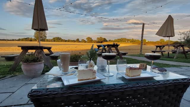 Desserts and drinks on a table with a scenic view of fields and trees under a sunset sky, patio umbrellas in the background.