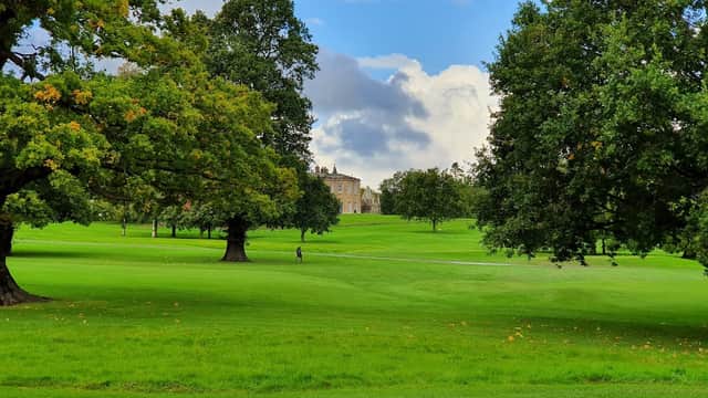 Lush green golf course with large trees and a historic building in the background under a partly cloudy sky.