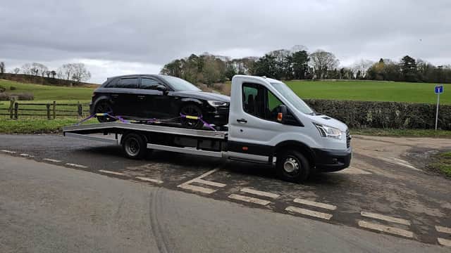 A flatbed truck with a black car secured on it, parked near a grassy field in Husthwaite, Yorkshire.