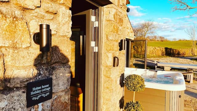 Stone cottage entrance with a sign, hot tub, and lush greenery under a bright blue sky.