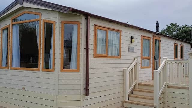 A holiday park cabin with large windows, a wooden deck, and green grass in Barmston, Yorkshire, under a cloudy sky.