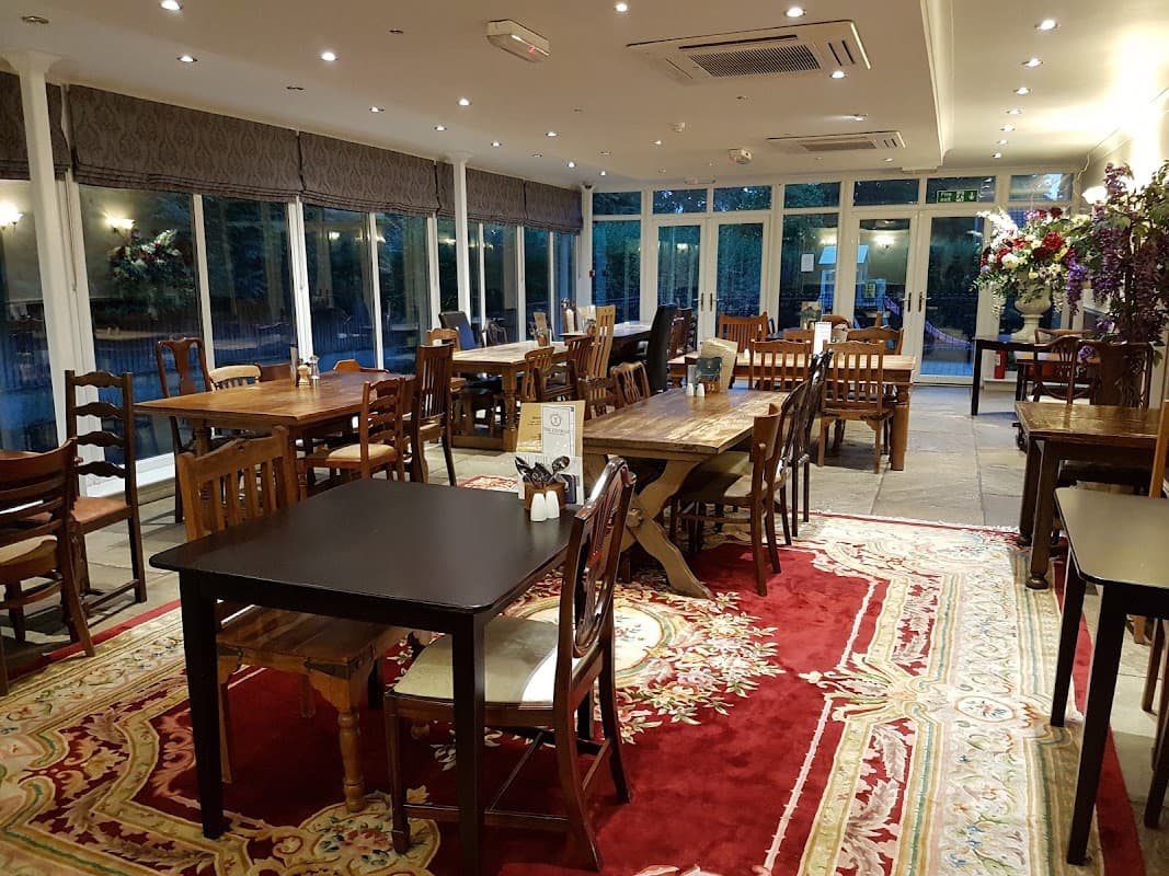 Cozy dining area with wooden tables, chairs, and large windows, featuring a patterned rug and floral decorations.