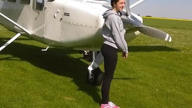 A woman stands beside a small white aircraft on a grassy field under a partly cloudy sky.