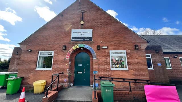 Brick building with a green door, welcoming sign, and colorful bins outside under a blue sky with clouds.