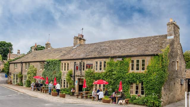 Historic stone pub with outdoor seating, red umbrellas, and patrons enjoying the scenic Yorkshire village.