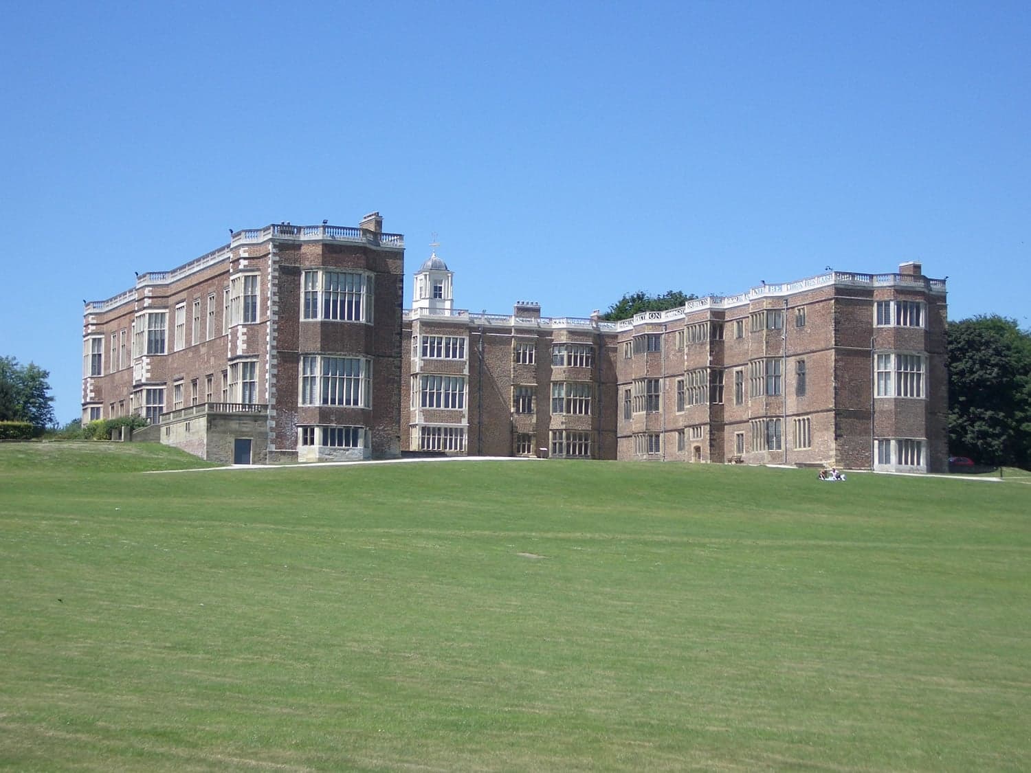 Historic brick mansion with large windows and expansive green lawn under a clear blue sky.