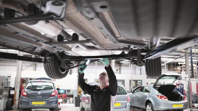 Mechanic inspecting a car's underside with a flashlight in a garage, surrounded by vehicles and workshop equipment.
