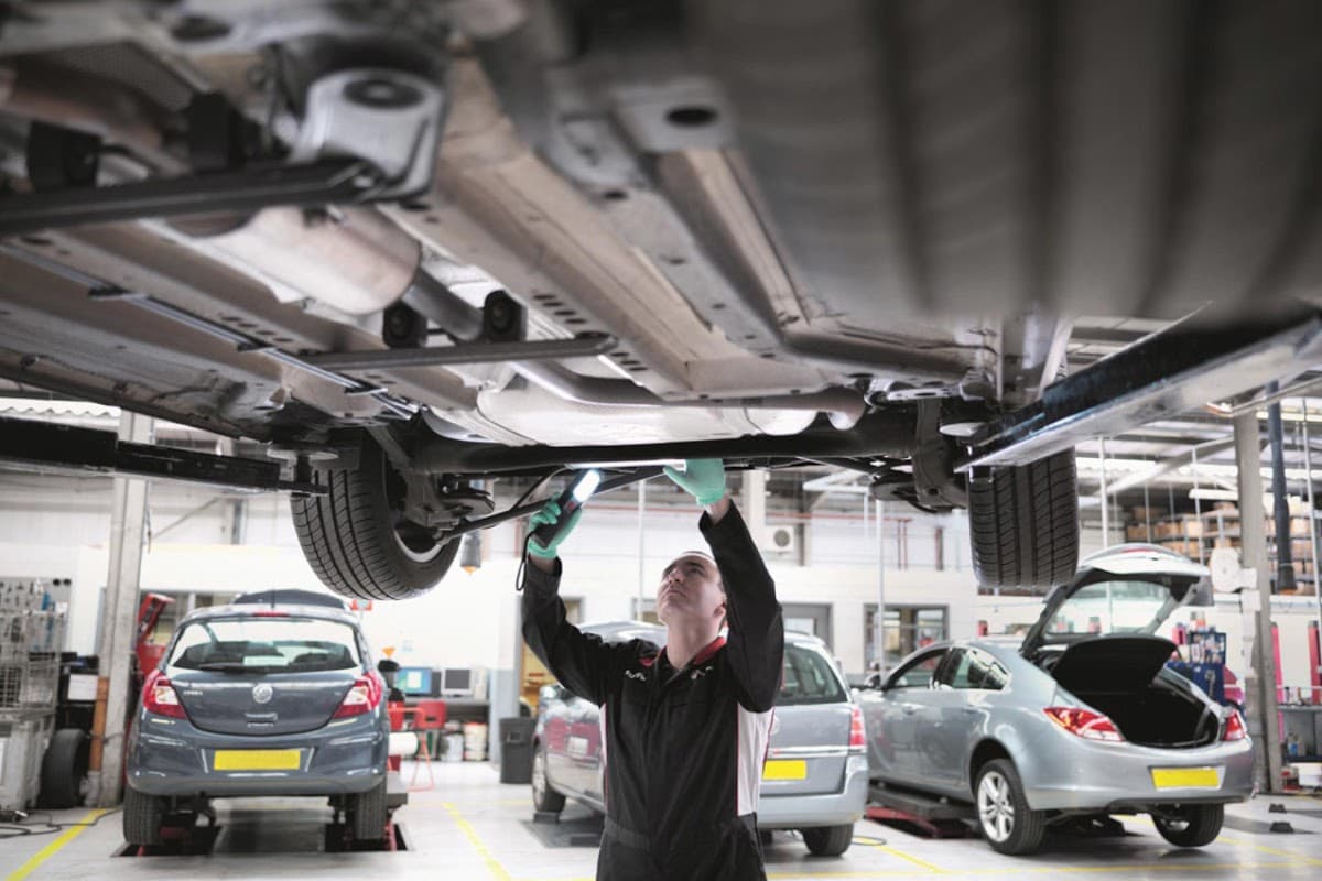 Mechanic inspecting a car's underside with a flashlight in a garage, surrounded by vehicles and workshop equipment.