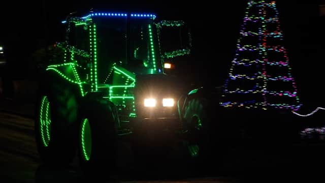 A brightly lit tractor adorned with colorful lights drives past a decorated Christmas tree at night.