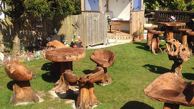 Wooden furniture in a grassy area, with carved tables and chairs, surrounded by greenery and a small shed.
