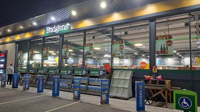 Budgens store front at night with illuminated signage, displays, and parking area. Greggs signage visible.