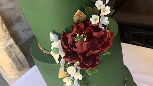 Three-tiered green cake adorned with a large burgundy flower, delicate white blossoms, and gold accents.