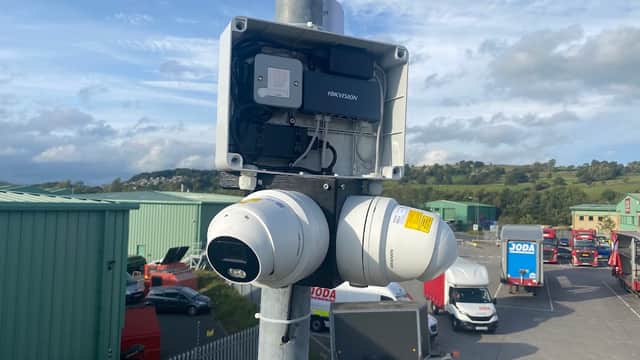 Surveillance cameras mounted on a pole, overlooking a parking lot with vehicles and green industrial buildings in the background.