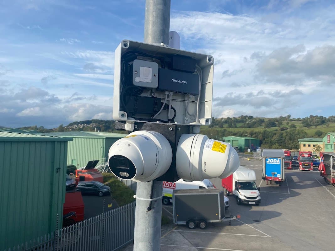 Surveillance cameras mounted on a pole, overlooking a parking lot with vehicles and green industrial buildings in the background.