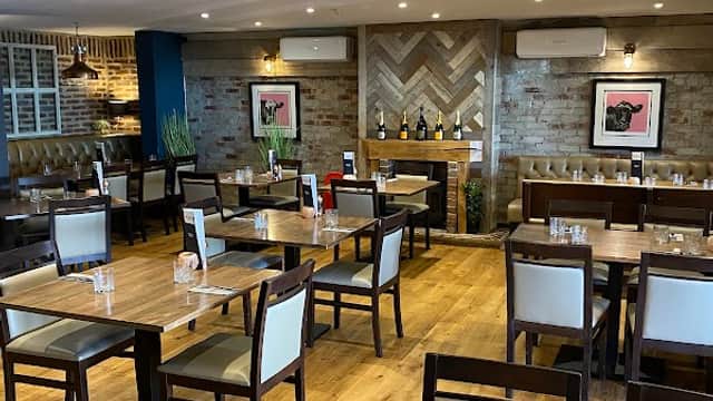 Cozy restaurant interior with wooden tables, chairs, and a stylish bar area featuring wine bottles and artwork.
