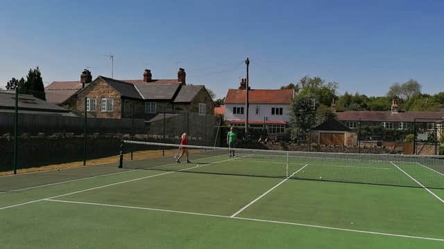 Tennis court with two players, surrounded by houses and trees under a clear blue sky in Collingham, Yorkshire.