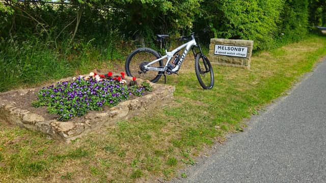 A bicycle rests beside a flower bed with colorful blooms and a sign reading "MELSONBY, a good place to live."