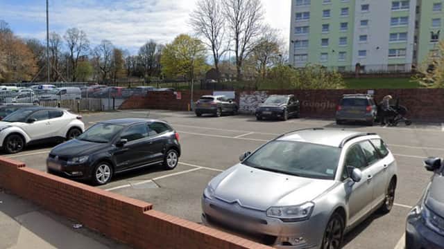Cars parked in a lot near a residential building, with trees and a clear sky in the background.