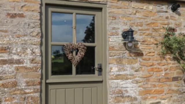 A rustic stone barn with a green door featuring a heart-shaped wreath and a light fixture beside it.