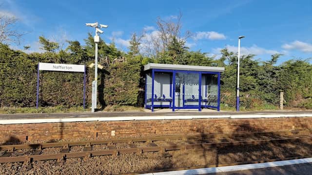 Nafferton train station with a blue shelter, sign, and overgrown greenery under a clear blue sky.