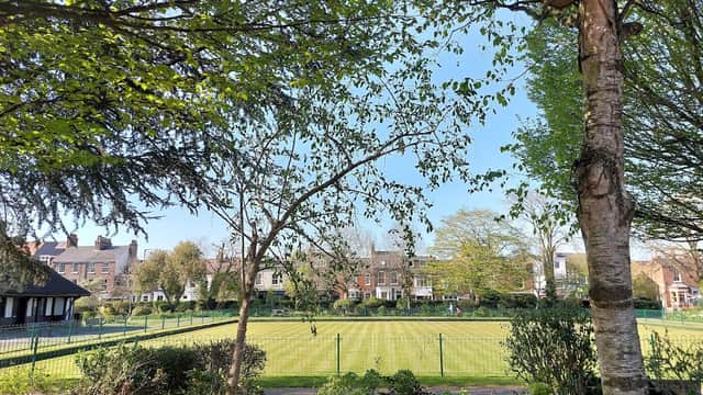 Lush green play area with trees, a manicured lawn, and residential buildings in the background under a clear blue sky.