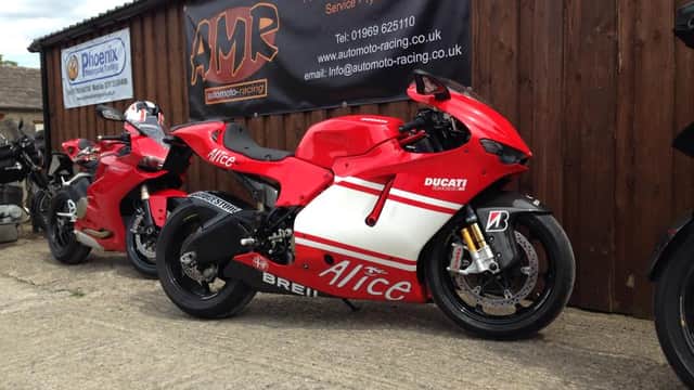 Red Ducati motorcycle with "Alice" branding parked outside a garage, alongside another bike, against a wooden wall.