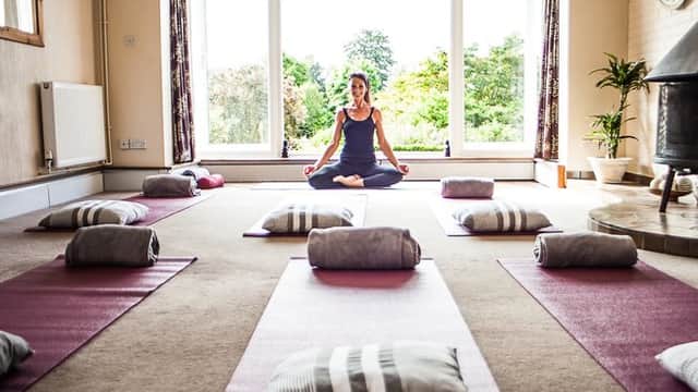 Yoga studio with mats and cushions arranged neatly, a woman meditating in a serene, sunlit space surrounded by greenery.
