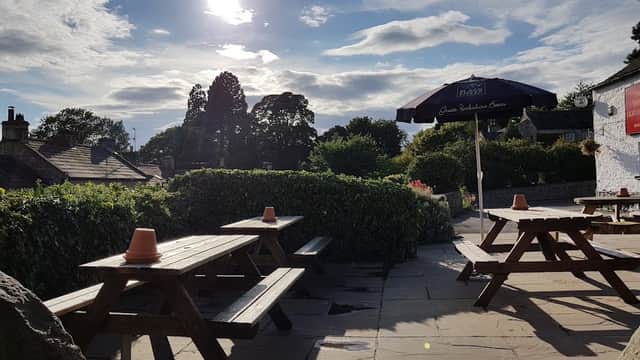 Outdoor seating area with wooden picnic tables, surrounded by greenery, under a sunny sky with clouds.