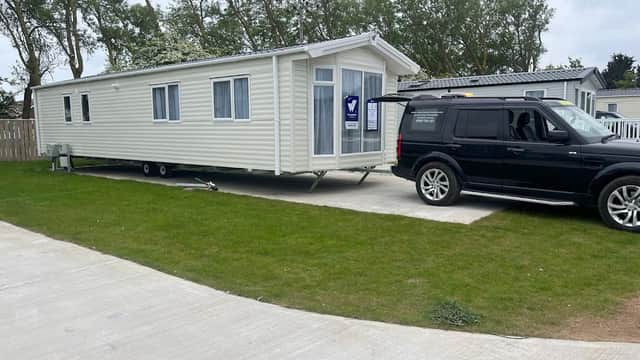 A static caravan parked on a concrete pad with a black SUV nearby, surrounded by green grass and trees.