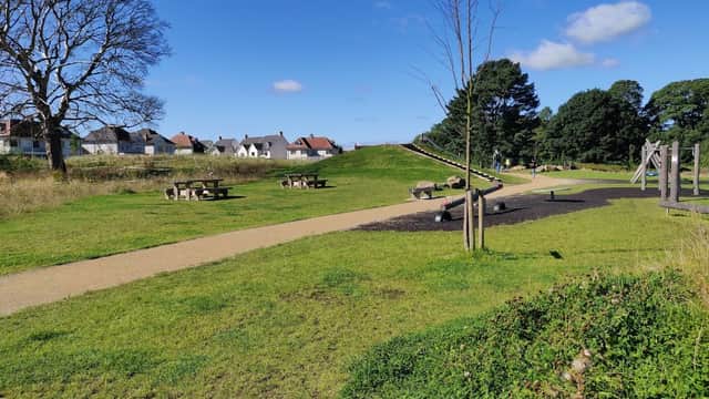 Lush green park with pathways, picnic tables, trees, and residential buildings under a clear blue sky.