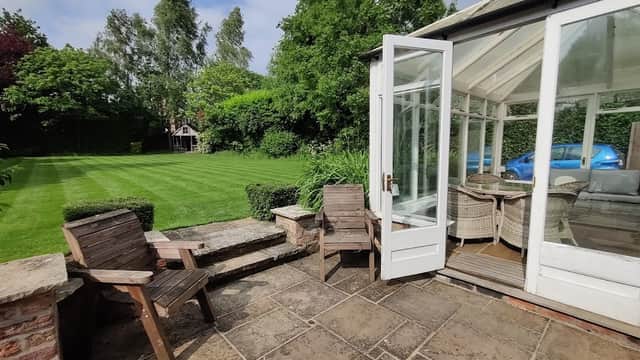 Sunlit patio area with wooden chairs, a glass conservatory, and a lush green garden in Appleton Roebuck, Yorkshire.