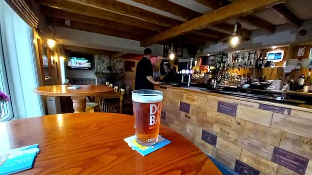 A pint of amber beer sits on a wooden table, with a rustic bar and bartender in the background.