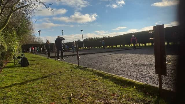 Petanque players on a gravel court under a bright sky, surrounded by greenery and wooden posts.