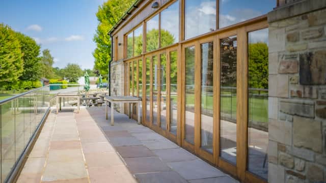 Wooden-framed glass doors open to a patio area with stone tiles, surrounded by lush green trees and a clear blue sky.