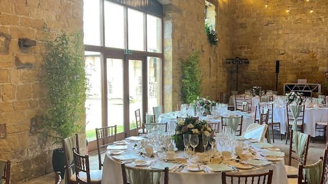 Elegant dining setup with round tables, white tablecloths, and greenery in a rustic stone venue with hanging lights.
