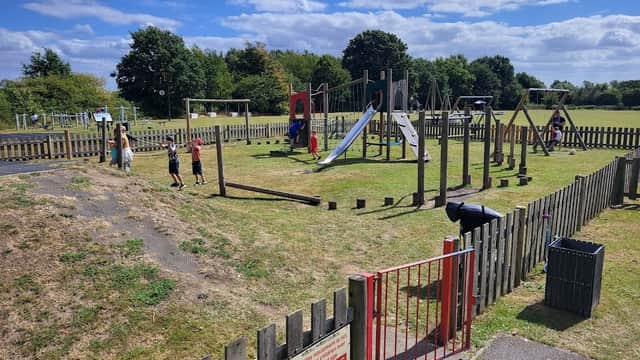 Children playing on various playground equipment in a sunny park with green grass and wooden fencing.
