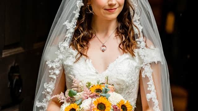 A bride in a lace dress and veil holds a vibrant bouquet of sunflowers and pastel flowers, smiling softly.