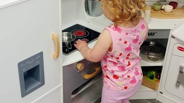 A toddler with curly hair plays at a toy kitchen, pretending to cook with various colorful food items around.