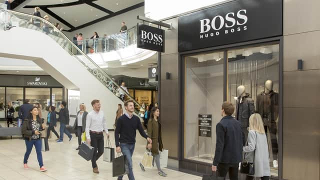 Shoppers with bags walk past Hugo Boss store in a modern, bright shopping mall with escalator and glass railings.