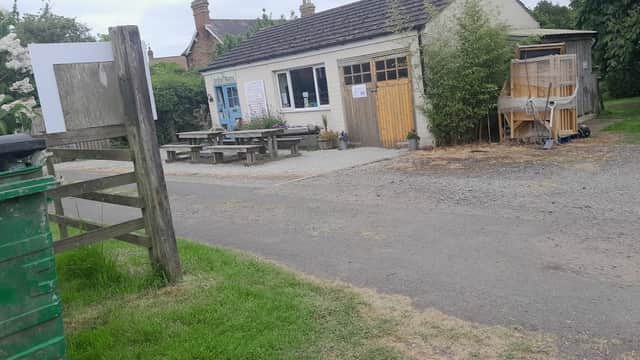 Campsite entrance with a building, picnic tables, green bin, and gravel path surrounded by trees and greenery.
