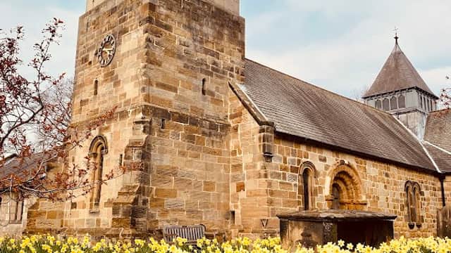 Sowerby Parochial Hall surrounded by blooming daffodils under a cloudy sky, featuring a stone tower and arched windows.