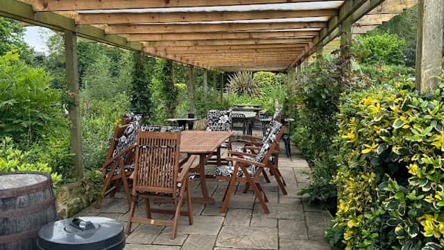 Outdoor dining area with wooden tables and chairs, surrounded by lush greenery and stone pathways.