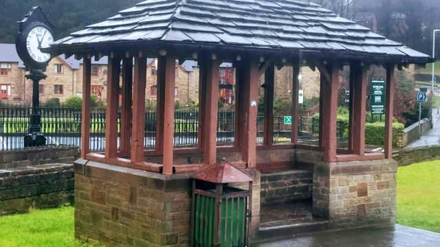 A wooden gazebo with a slate roof, next to a clock and a green waste bin, in a grassy area of a car park.