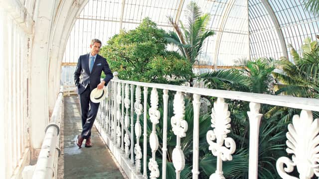Man in a suit holding a hat, walking on a balcony inside a glass conservatory with lush green plants.