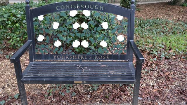 Black bench with white roses and "Conisbrough Yorkshire's Rose" engraved, surrounded by greenery and fallen leaves.