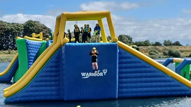 Colorful inflatable water obstacle course with people in helmets, jumping and swimming in a lake under a clear blue sky.