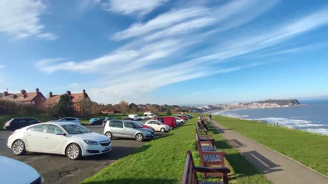 Free parking area along a coastal road with cars, benches, and a view of the sea and distant town under a blue sky.